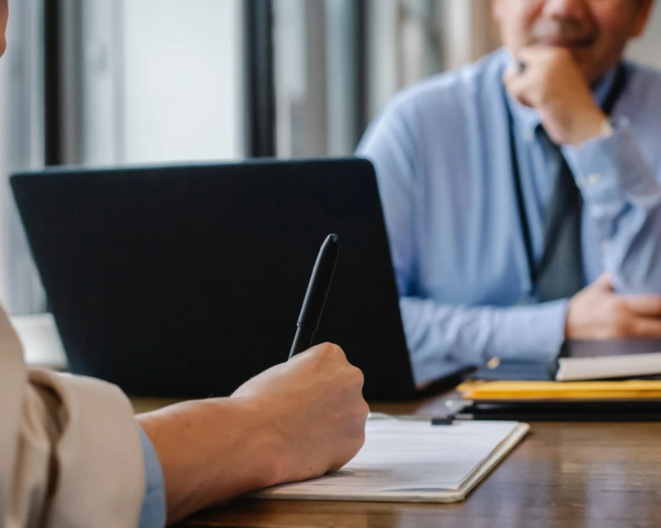 Student interviews for university admissions: A hand holds a pen, writing on a clipboard, while the interviewer attentively evaluates the applicant's profile with folded hands and a thoughtful expression.